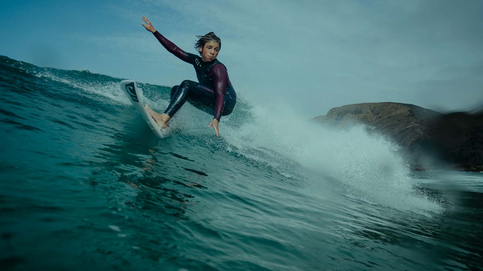 Young Ripper surfing a wave. Full Wetsuit with Dark Red sleeves. Glassy Water Conditions.