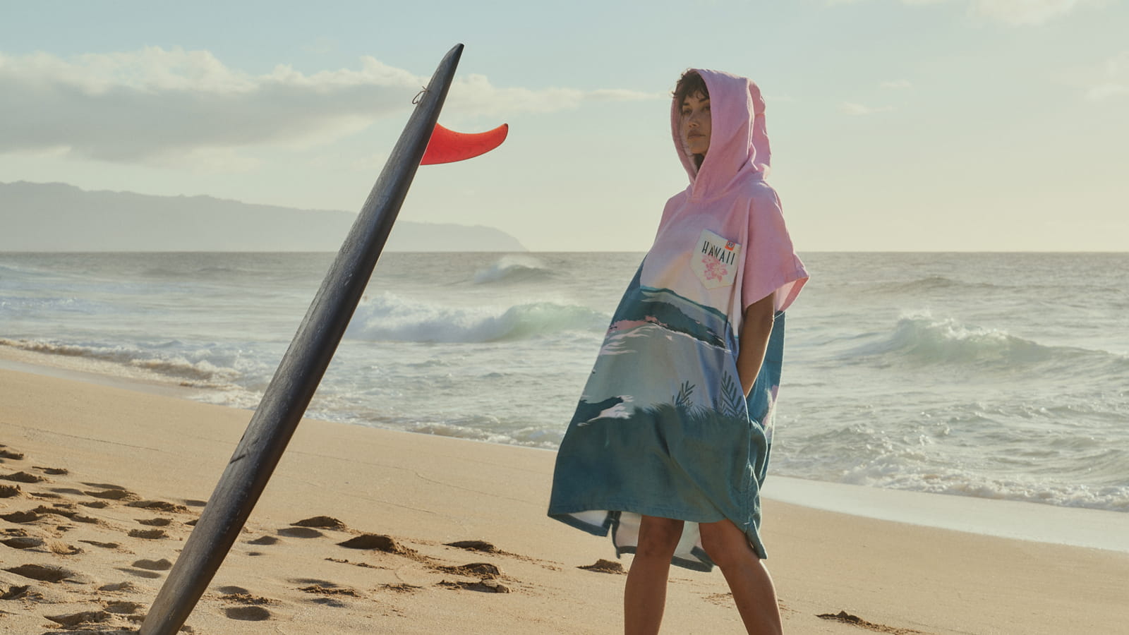 Girl in a bright cozy poncho on the beach near a surfboard.