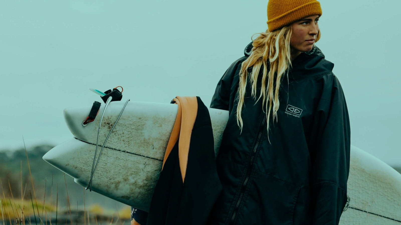 Blond Girl in Jacket Poncho holding in hand surfboard and wetsuit.