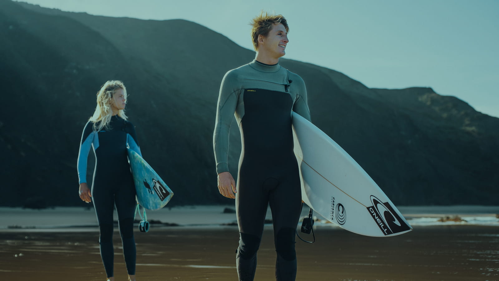 Mens Wetsuits. Male in a full wetsuits standing in front and female at the background. Surfboards in hands, ready to go surfing.